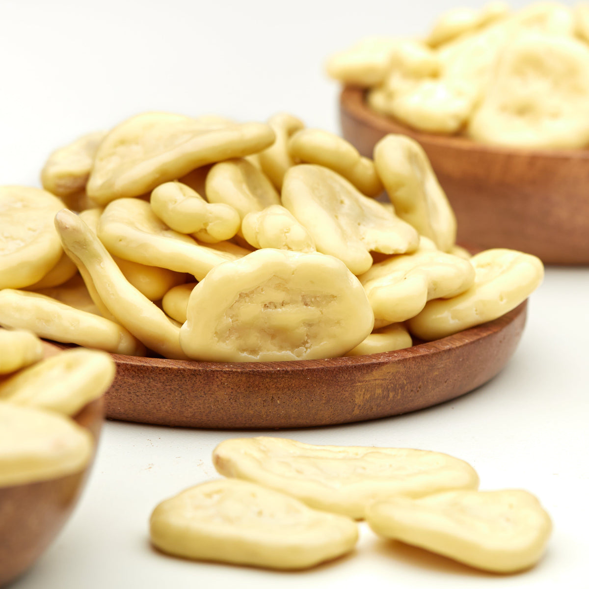 Knusprige Bananenchips in weißer Schokolade von NESSLIM auf einem Holzteller, mit einzelnen Chips im Vordergrund.
