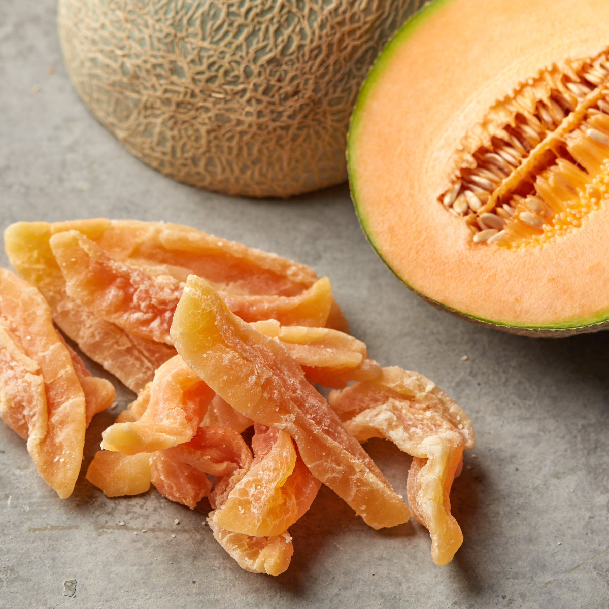 Getrocknete Cantaloupe-Melonenstreifen auf grauem Untergrund neben einer ganzen und einer halbierten Melone mit orangefarbenem Fruchtfleisch.