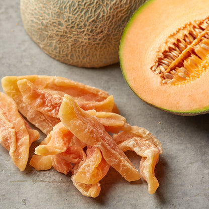 Getrocknete Cantaloupe-Melonenstreifen auf grauem Untergrund neben einer ganzen und einer halbierten Melone mit orangefarbenem Fruchtfleisch.