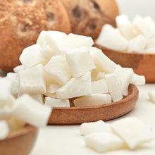 Süße Jumbo-Kokoswürfel von NESSLIM als tropischer Kokos-Snack, auf einem Holzteller neben ganzen Kokosnüssen angerichtet.