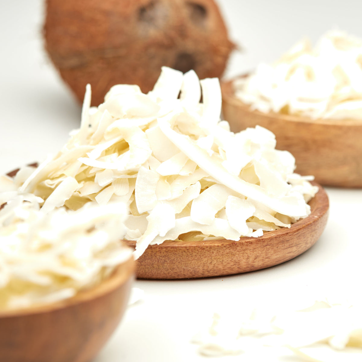 Weiße Kokoschips von NESSLIM als knuspriges Topping für Bowl, Müsli und Joghurt, mit einer ganzen Kokosnuss im Hintergrund.